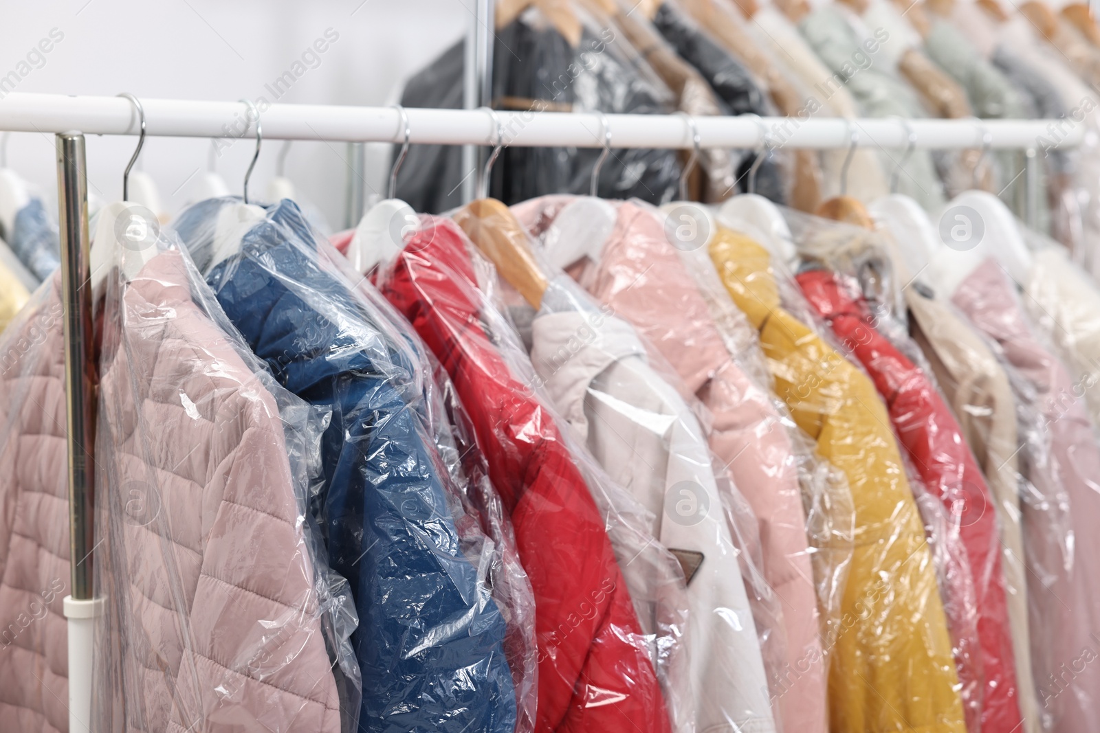 Photo of Dry-cleaning service. Many different clothes in plastic bags hanging on rack indoors, closeup