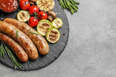 Photo of Slate plate with delicious barbecued sausages and vegetables on gray background. Space for text
