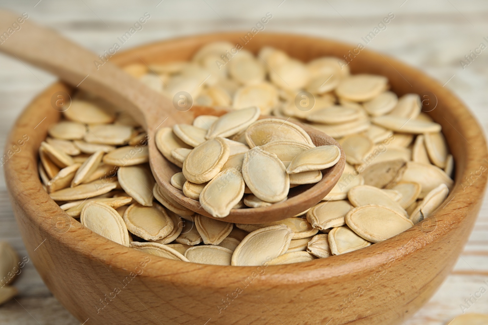 Photo of Wooden bowl and spoon of raw pumpkin seeds on white table, closeup