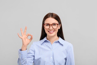 Portrait of smiling woman in stylish eyeglasses showing ok gesture on grey background