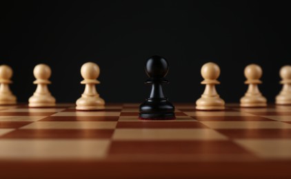 Photo of Black pawn in front of white ones on wooden chess board against dark background