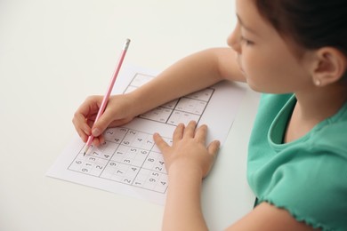 Photo of Little girl solving sudoku puzzle at white table, closeup