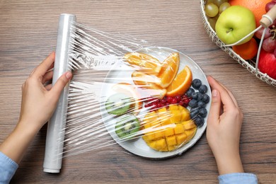 Photo of Woman putting plastic food wrap over plate of fresh fruits and berries at wooden table, top view