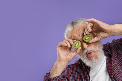 Photo of Funny senior man covering eyes with halves of kiwi on purple background, space for text