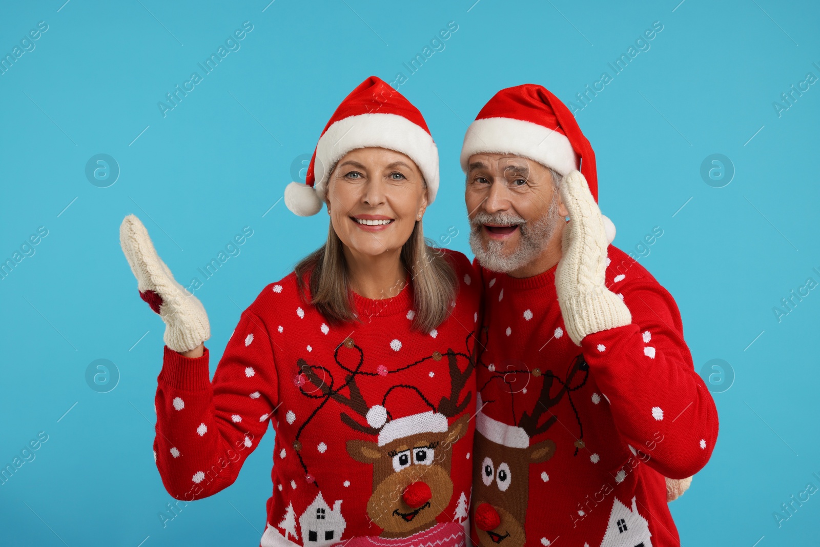 Photo of Happy senior couple in Christmas sweaters, Santa hats and knitted mittens on light blue background