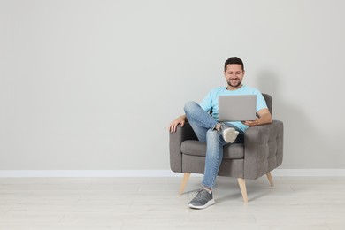 Happy man sitting in armchair and using laptop indoors, space for text