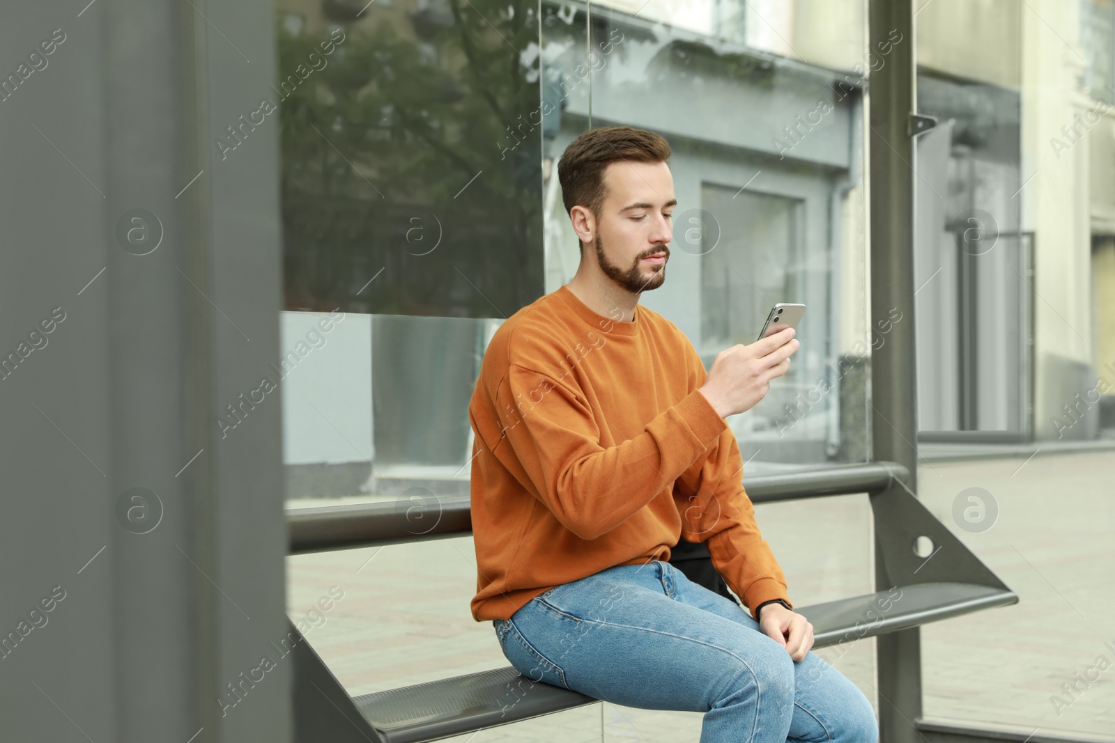 Photo of Young man with smartphone waiting for public transport at bus stop