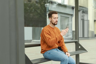 Photo of Young man with smartphone waiting for public transport at bus stop