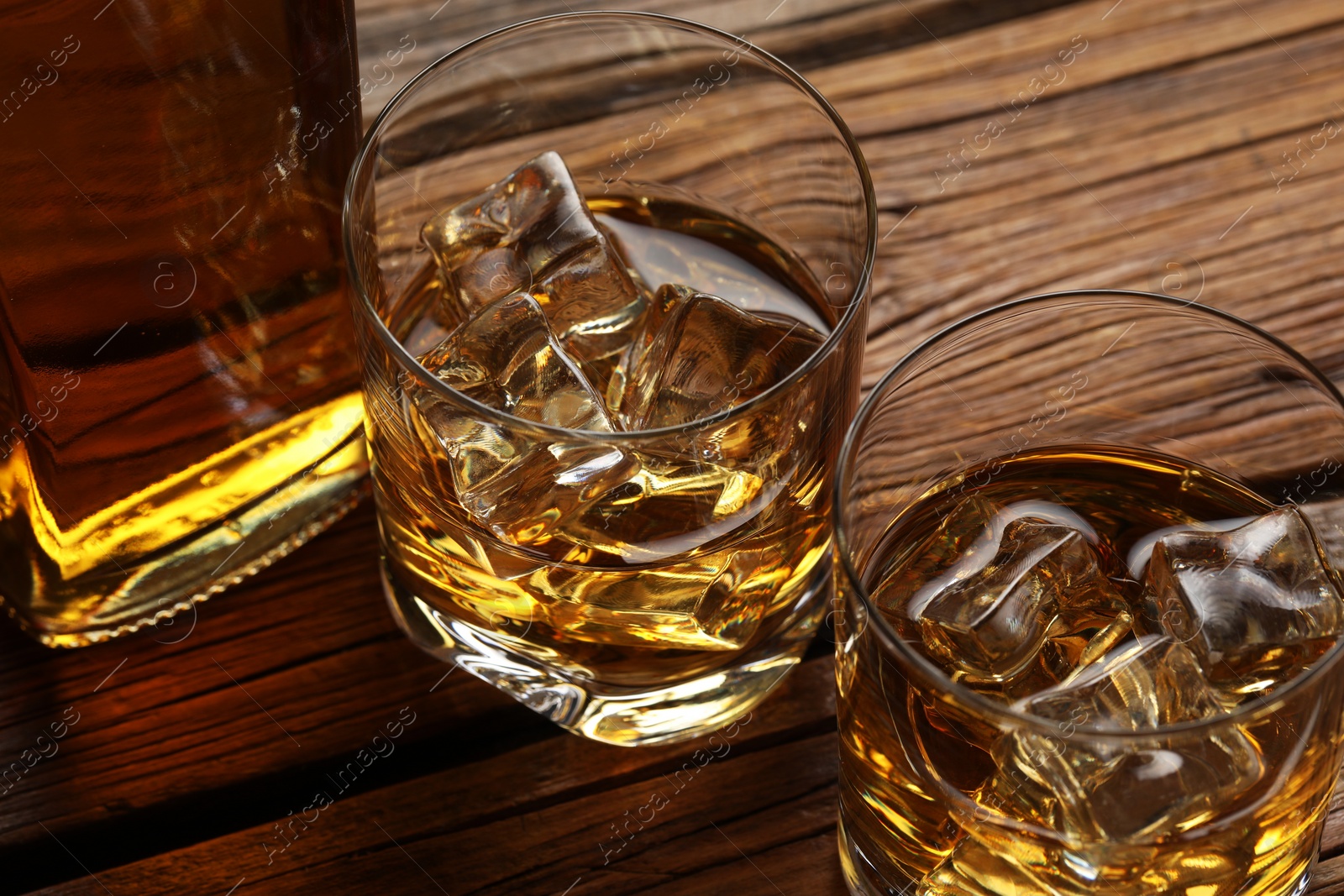 Photo of Whiskey with ice cubes in glasses and bottle on wooden table, closeup