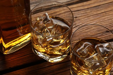 Whiskey with ice cubes in glasses and bottle on wooden table, closeup