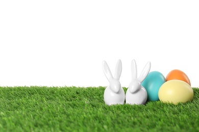 Photo of Ceramic Easter bunnies and dyed eggs on green grass against white background, space for text