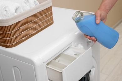 Man pouring fabric softener from bottle into washing machine indoors, closeup