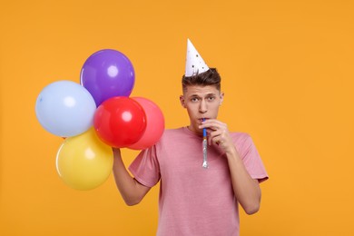Photo of Young man in party hat with bunch of balloons and blower on orange background