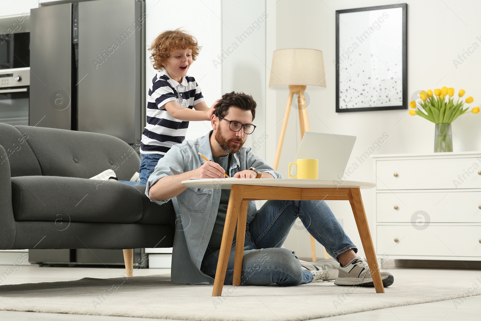 Photo of Little boy bothering his father at home. Man working remotely at desk