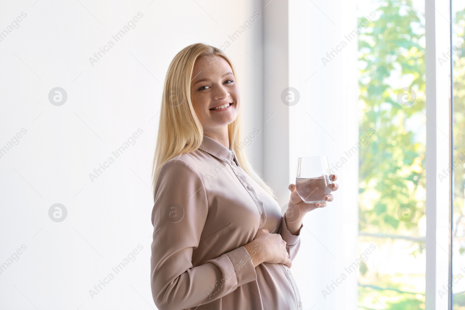 Photo of Young pregnant woman with glass of water near window indoors