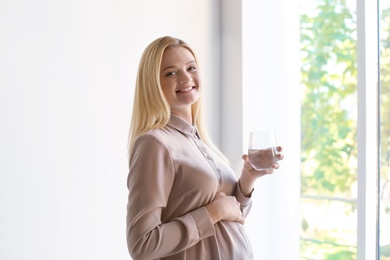 Young pregnant woman with glass of water near window indoors