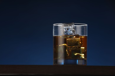 Photo of Whiskey with ice cubes in glass on table against dark blue background. Space for text