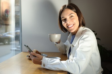 Special Promotion. Happy young woman with cup of drink using smartphone at table in cafe