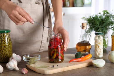 Photo of Woman taking pickled chili pepper from jar at wooden table indoors, closeup