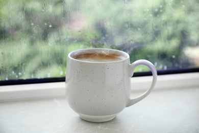 Cup of hot coffee near window on rainy day