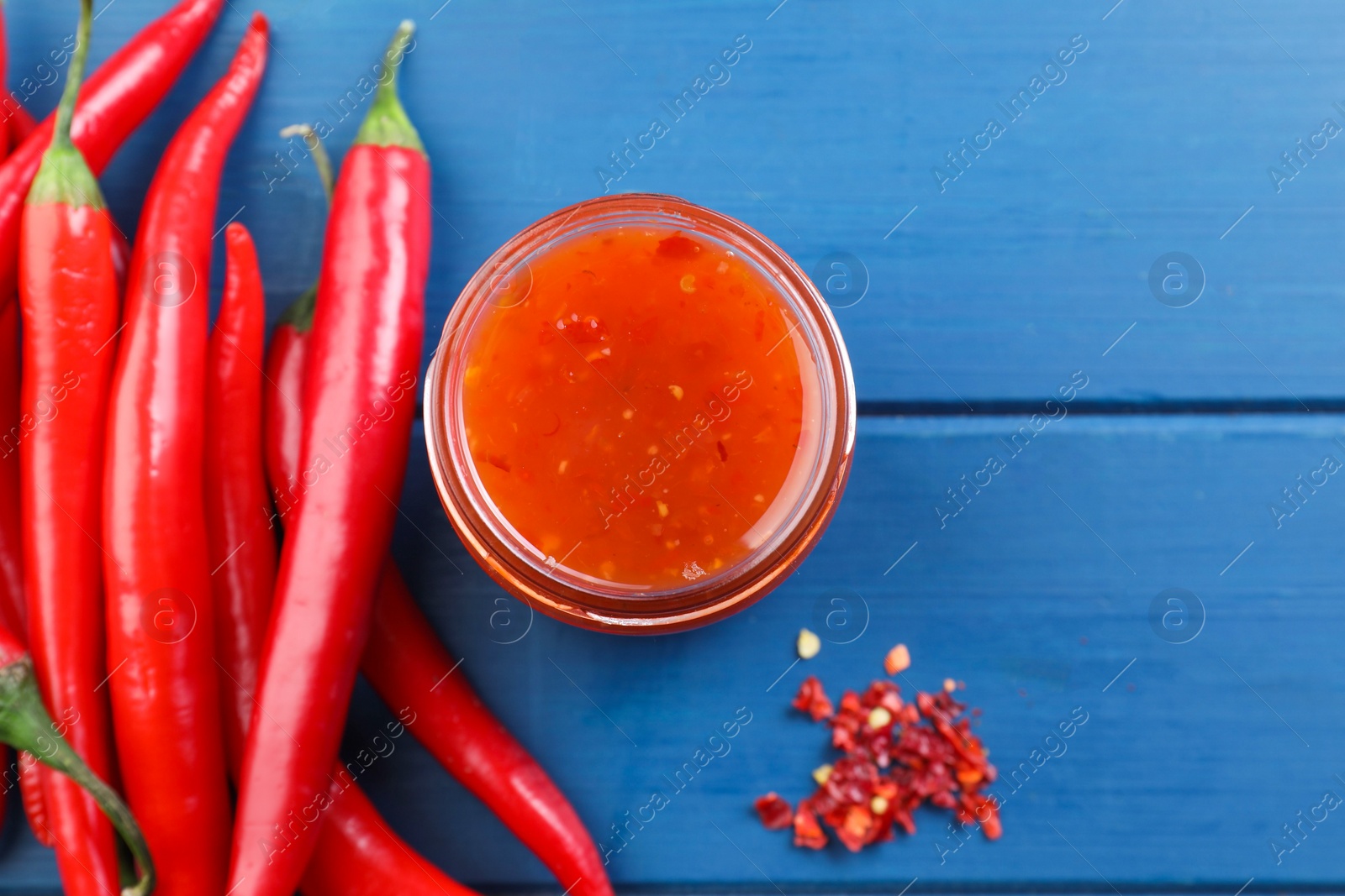 Photo of Spicy chili sauce in jar and peppers on blue wooden table, flat lay. Space for text