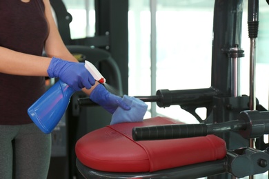 Photo of Woman cleaning exercise equipment with disinfectant spray and cloth in gym, closeup