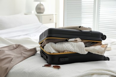 Photo of Open suitcase full of clothes, jacket and fashionable accessories on bed in room