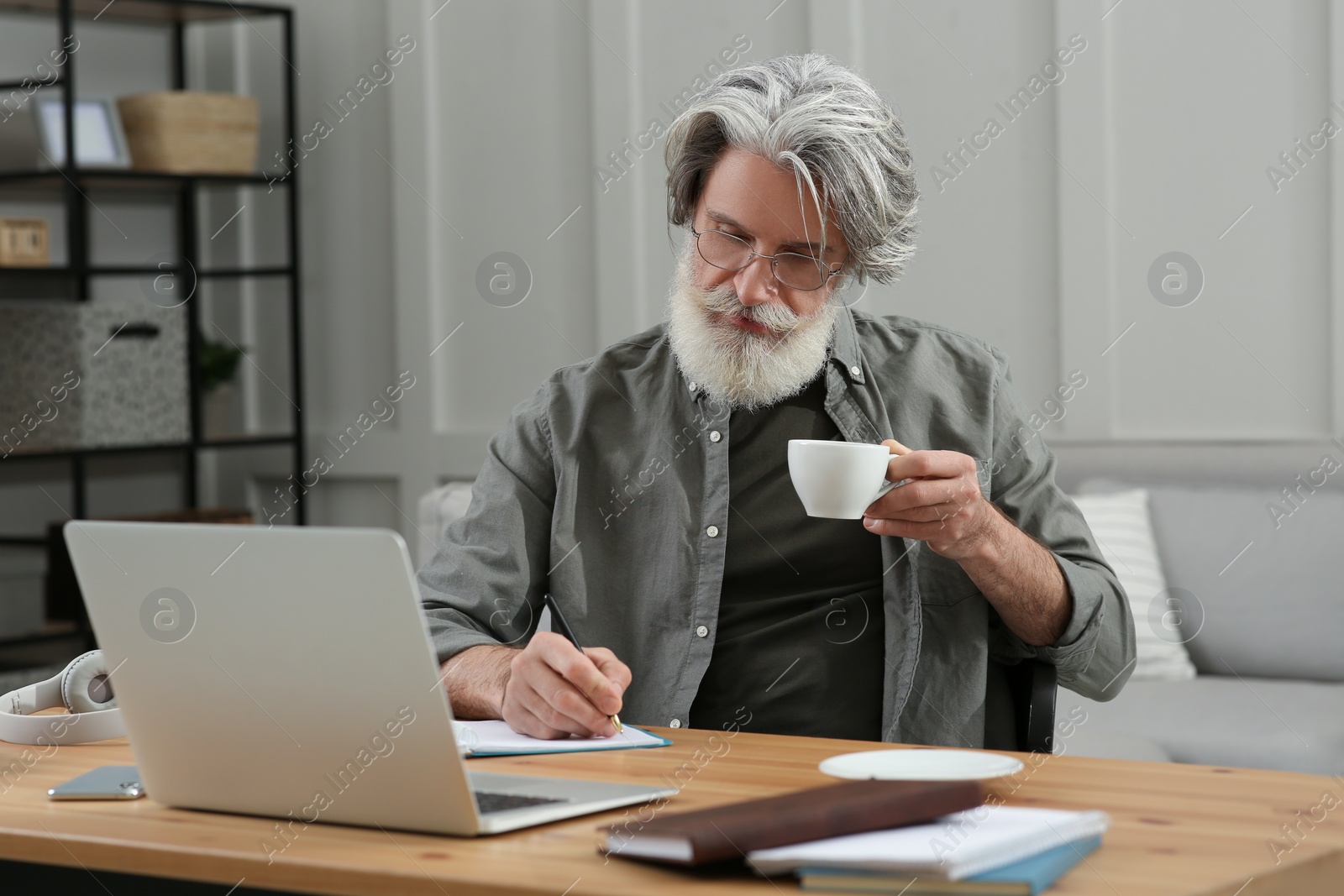 Photo of Middle aged man with laptop, notebook and cup of drink learning at table indoors