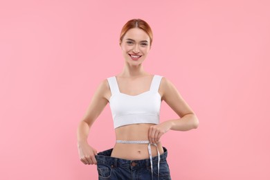 Photo of Slim woman wearing big jeans and measuring waist with tape on pink background. Weight loss