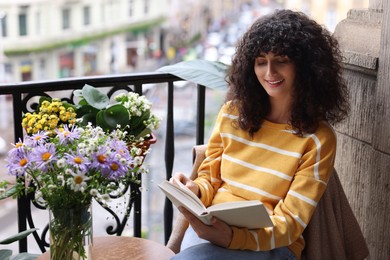 Young woman reading book at table with beautiful flowers on balcony