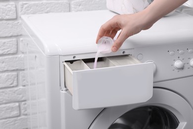 Photo of Woman pouring fabric softener from cap into washing machine near white brick wall, closeup