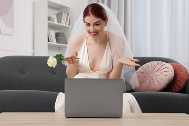 Photo of Happy bride with rose having online video chat via laptop in living room