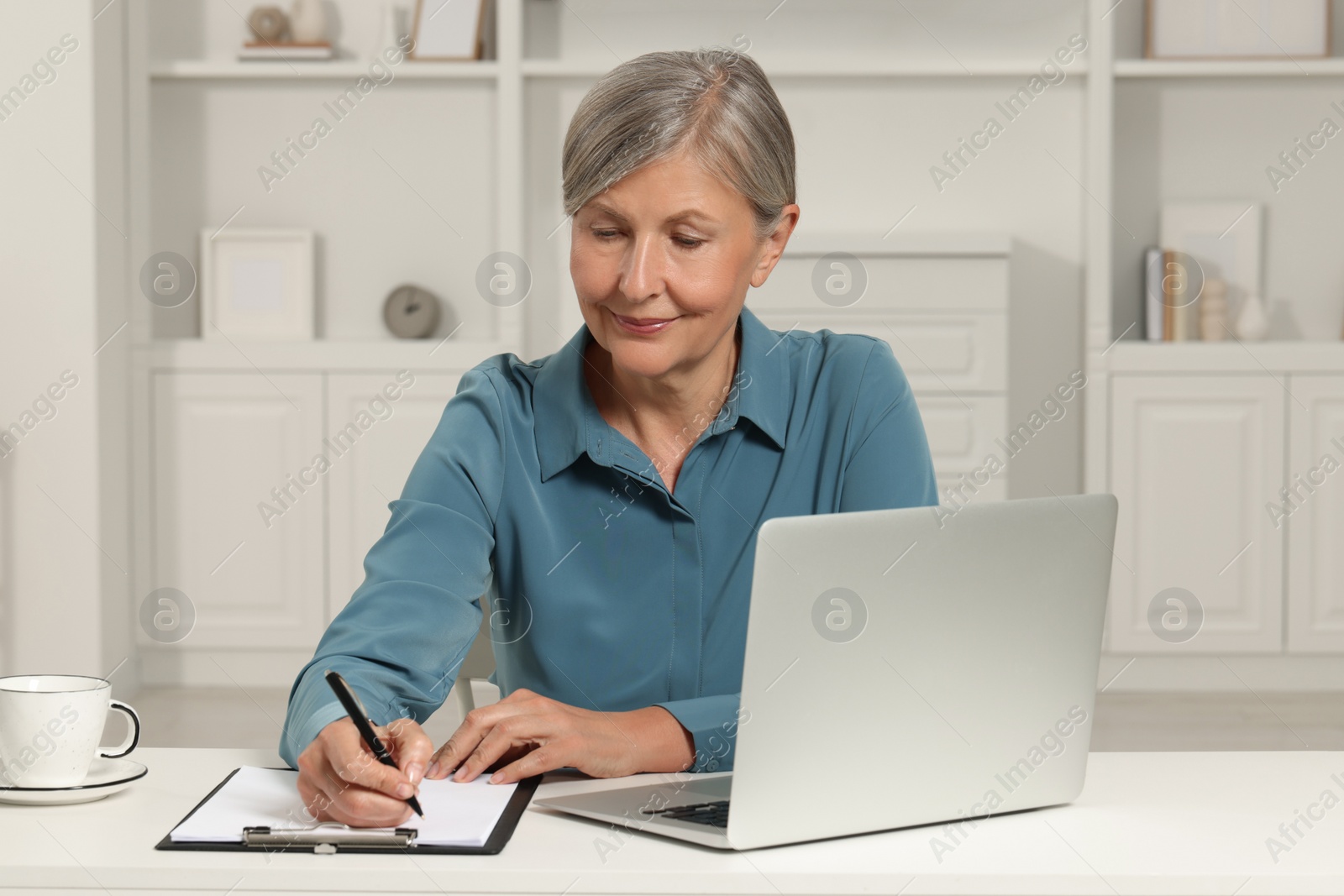 Photo of Beautiful senior woman taking notes near laptop at white table indoors