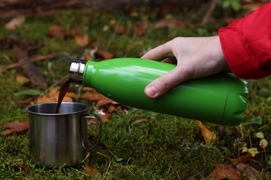 Photo of Woman pouring hot instant coffee from thermo bottle  into mug on green grass outdoors, closeup