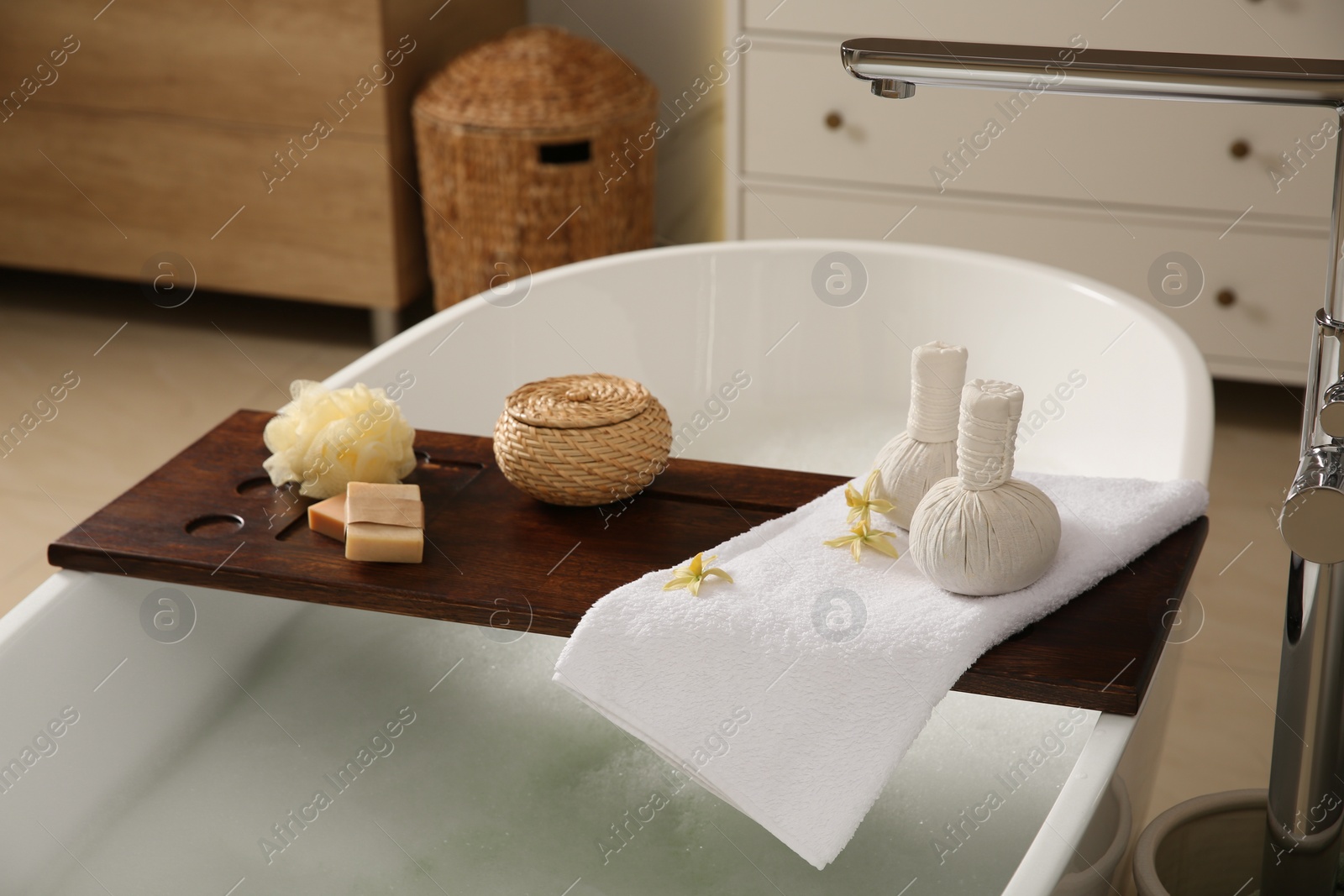 Photo of Wooden bath tray with herbal massage bags and bathroom amenities on tub indoors