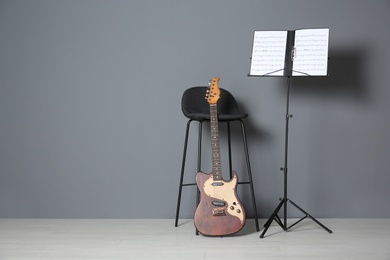 Photo of Electric guitar, chair and note stand with music sheets near grey wall indoors. Space for text