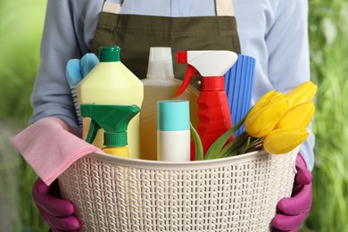 Photo of Woman holding basket with spring flowers and cleaning supplies outdoors, closeup