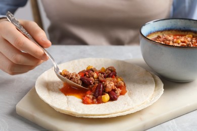 Photo of Woman putting tasty chili con carne into tortilla at white table, closeup