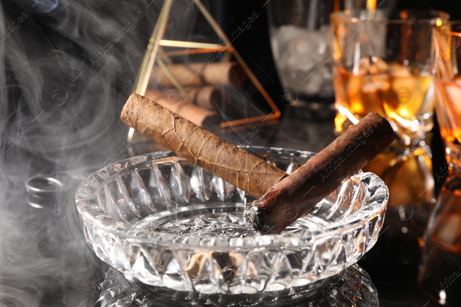 Photo of Smoldering cigars, ashtray and whiskey on black mirror surface, closeup