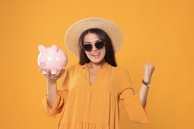Happy young woman in stylish sunglasses and straw hat with piggy bank on orange background