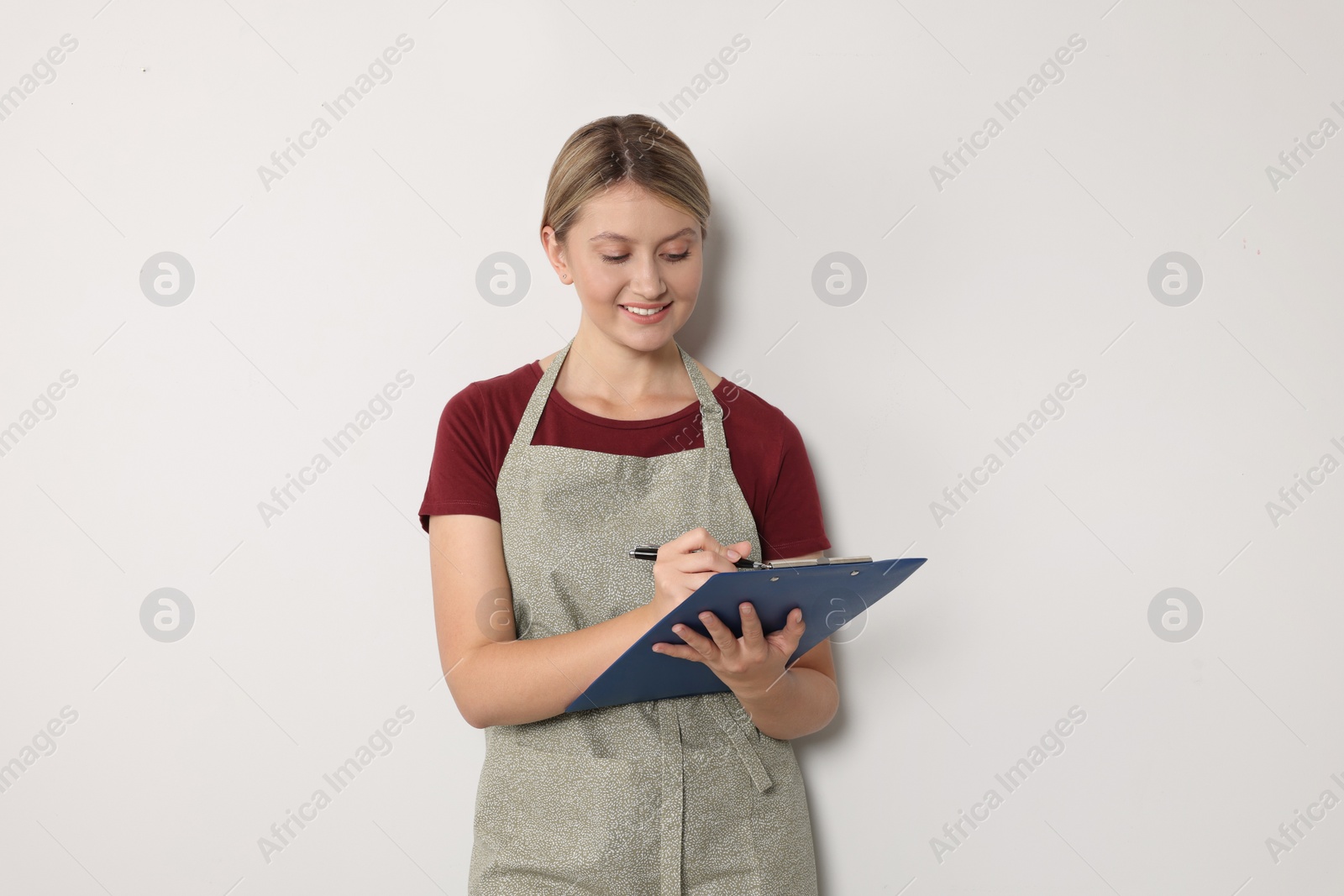 Photo of Beautiful young woman in clean apron with clipboard on light grey background