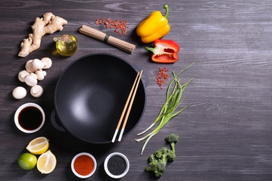 Photo of Empty iron wok and chopsticks surrounded by ingredients on dark grey wooden table, flat lay. Space for text