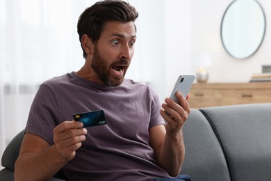 Photo of Shocked man with credit card using smartphone on sofa at home. Be careful - fraud