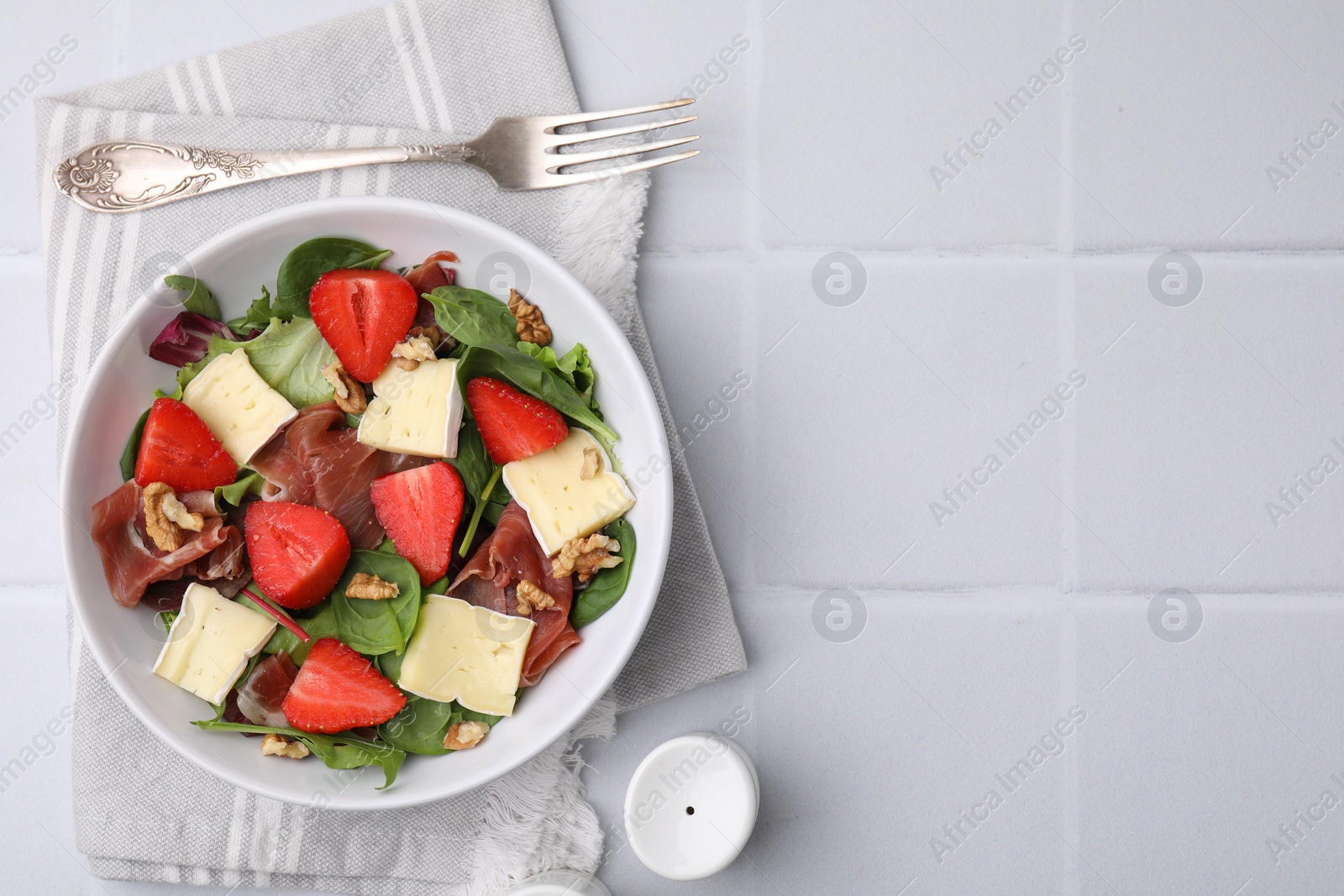 Photo of Tasty salad with brie cheese, prosciutto, strawberries and walnuts served on white tiled table, flat lay. Space for text