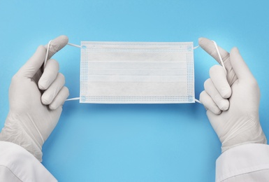 Photo of Doctor in medical gloves holding protective mask on light blue background, closeup