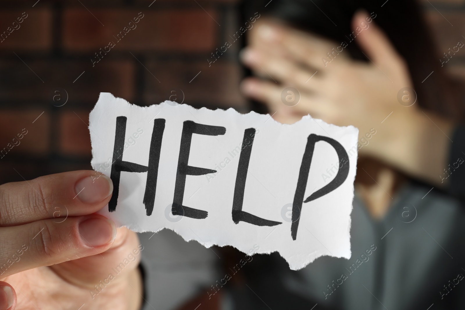 Photo of Domestic violence concept. Unhappy woman holding paper with written word Help near brick wall, focus on hand