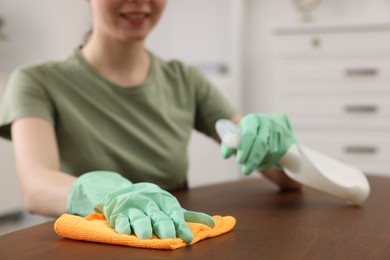 Photo of Woman with spray bottle and microfiber cloth cleaning wooden table in room, closeup