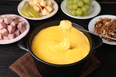 Dipping piece of ham into fondue pot with melted cheese on black wooden table, closeup