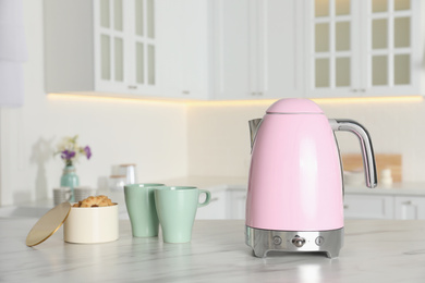 Modern electric kettle, cups and cookies on table in kitchen