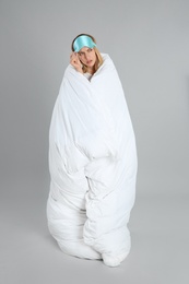 Emotional young woman in sleeping mask wrapped with soft blanket on grey background